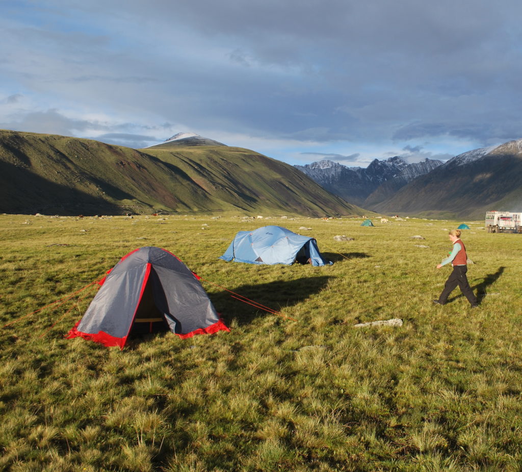 Morning at camp in a grassy valley. A woman walks through a group of three tents