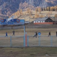 Children play soccer in a fenced simple field. In the background there are low buildings and a mountain slope rises behind them.