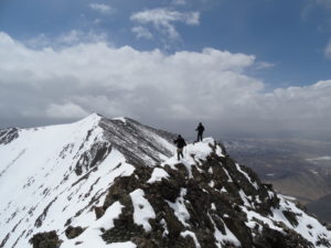 Two figure walk a narrow snow-covered ridge on a mountain top